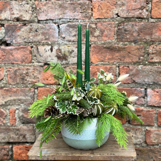 Twelfth Night Christmas table arrangement with green candles ivory roses pine cones waxflower holly and winter foliage displayed in a ceramic bowl.